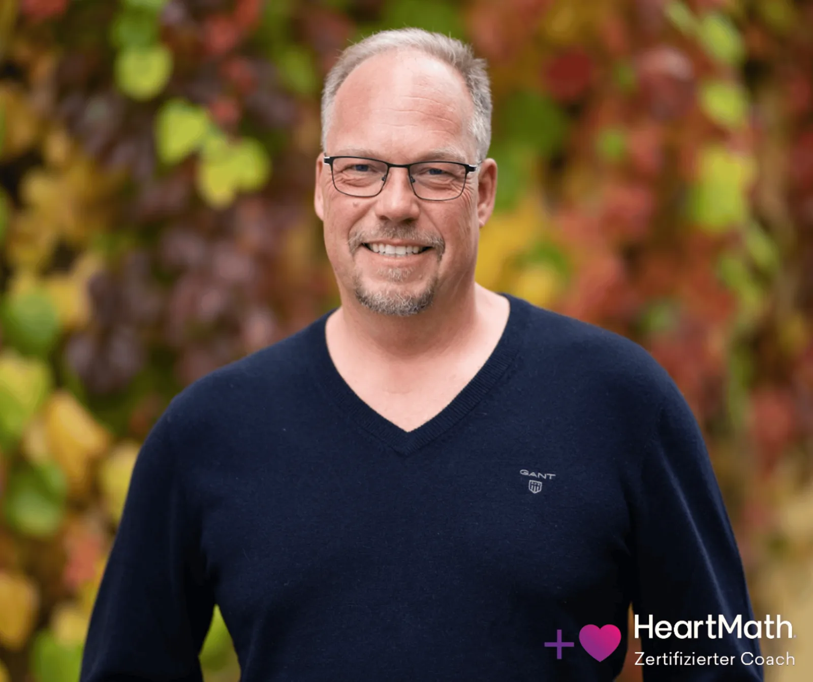 Florian Scherbauer, lächelnd, mit Brille und dunkelblauem Pullover vor Herbstlaub